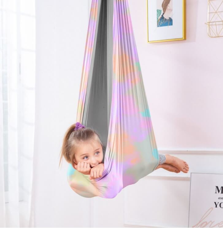 Kind liegt in einer mehrfarbigen Therapieschaukel mit pastellfarbenem Regenbogenmuster und grauer Innenseite in einem hellen Zimmer.