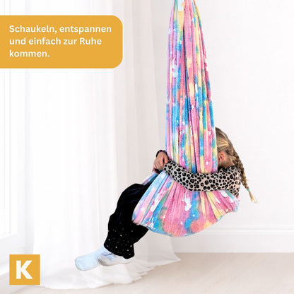 Kind in der Glow Therapieschaukel Pink Horse beim Schaukeln in buntem Stoff mit Einhornmuster
