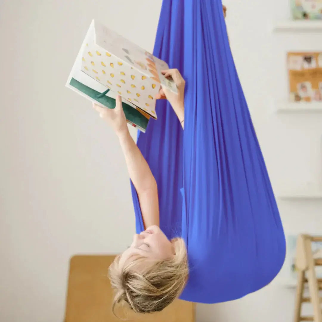 Kind liegt entspannt in einer blauen Therapieschaukel und liest ein Buch in einem hellen Kinderzimmer.