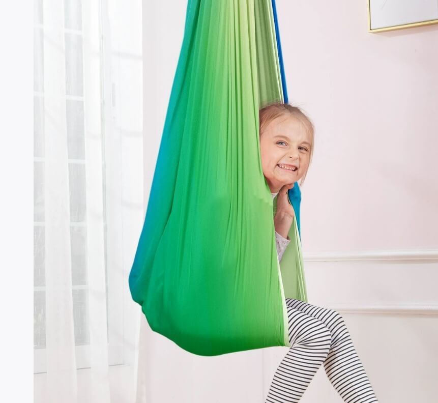 Kind sitzt lachend in einer grün-blauen Therapieschaukel in einem hellen Zimmer.