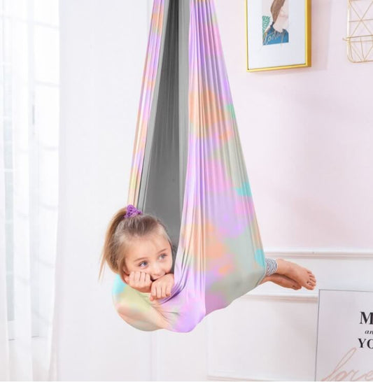 Kind liegt in einer mehrfarbigen Therapieschaukel mit pastellfarbenem Regenbogenmuster und grauer Innenseite in einem hellen Zimmer.