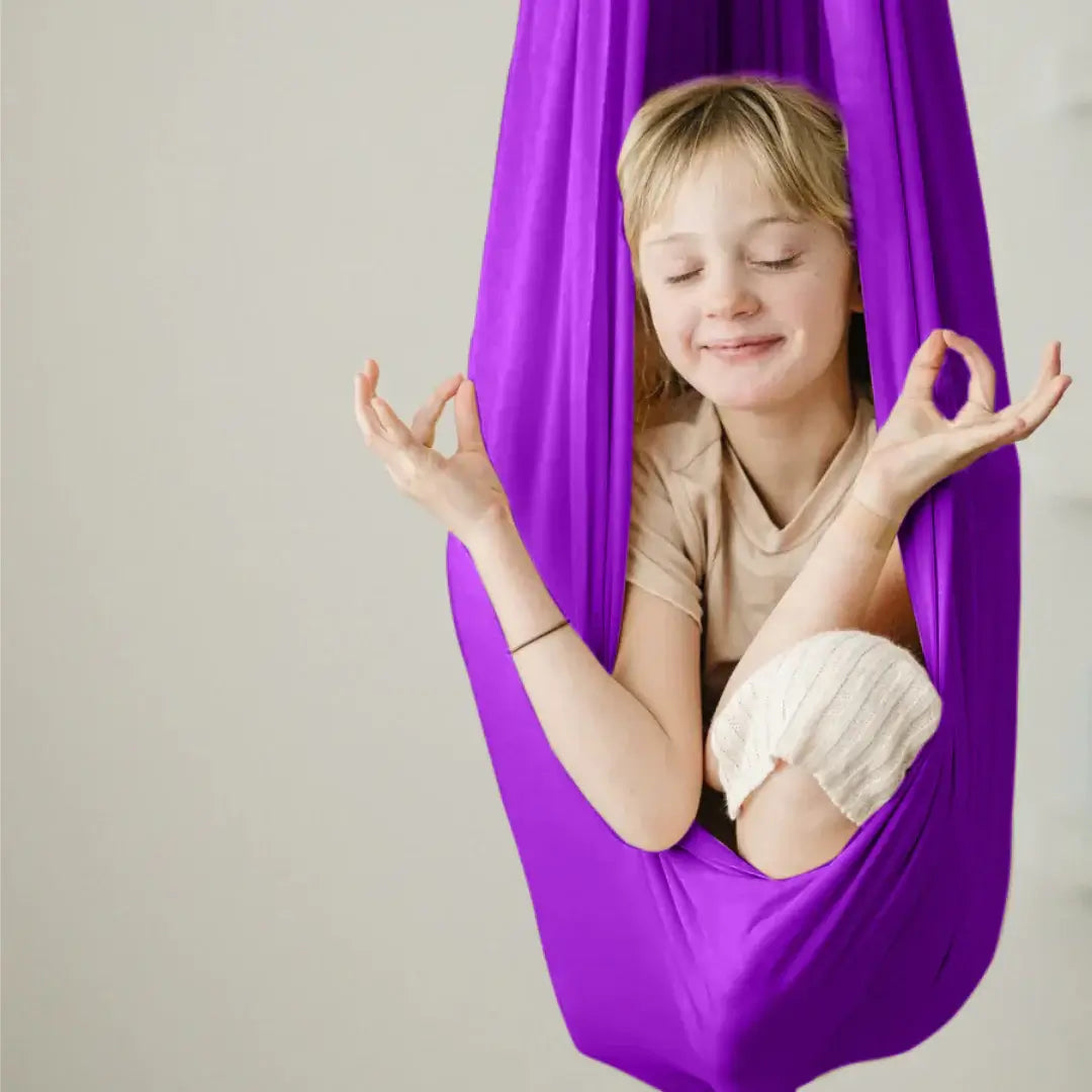 Mädchen sitzt entspannt in einer violetten Therapieschaukel und macht eine Meditationspose mit geschlossenen Augen.
